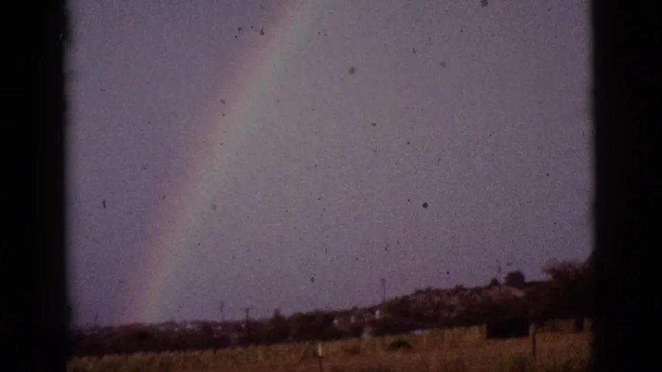 1965 8mm Tucson-area frame with a rainbow over open ground, low vegetation, fence posts, and visible dark film borders.