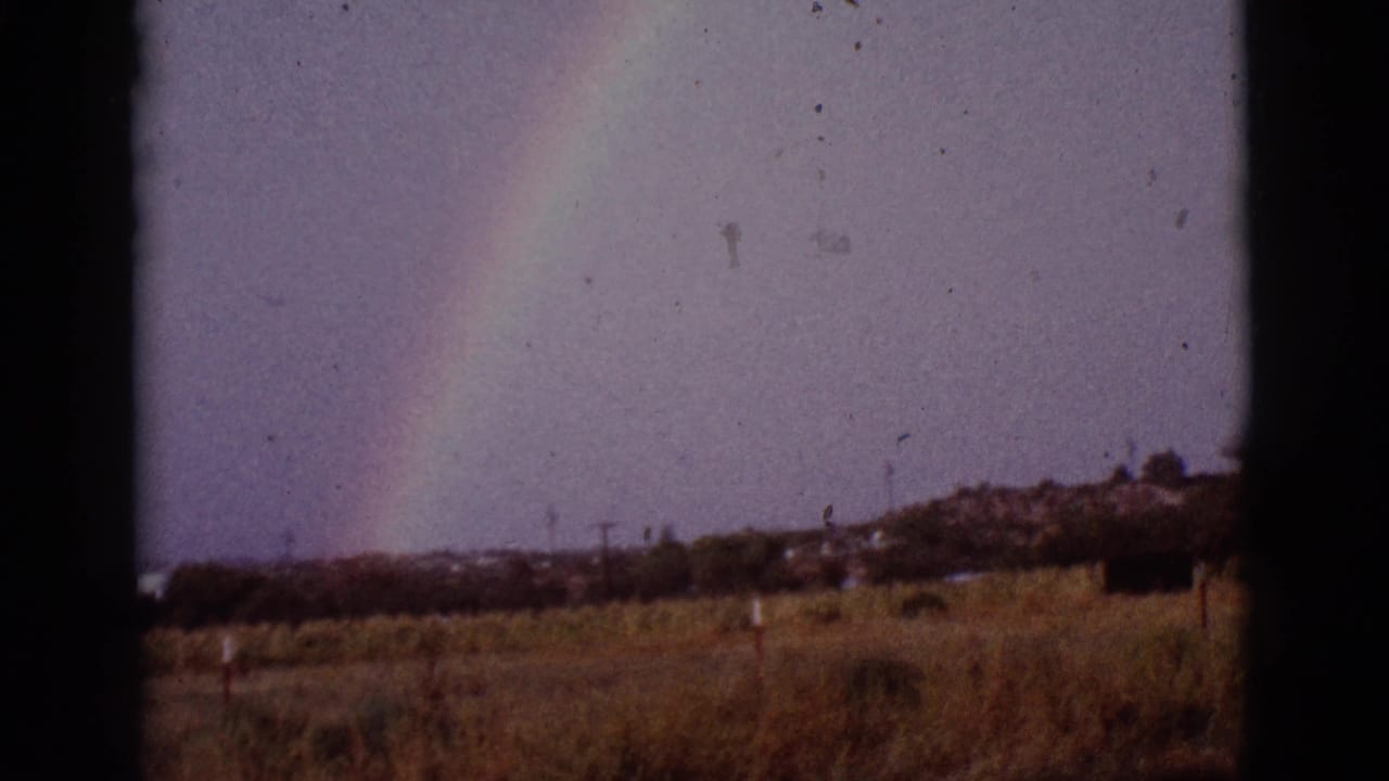 Rainbow over open field with distant trees and low structures.