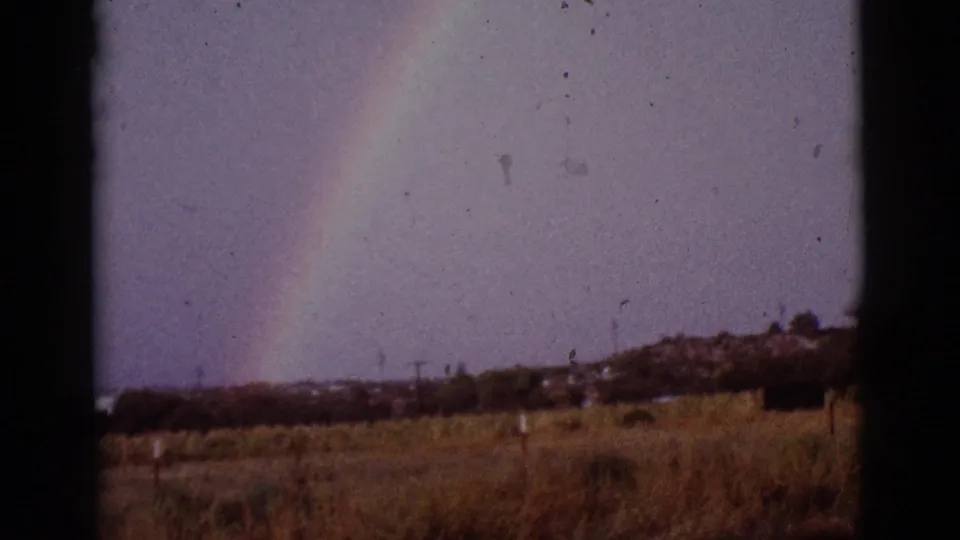 Rainbow remains visible above a dry Tucson-area field, with scattered distant trees or structures along the horizon.