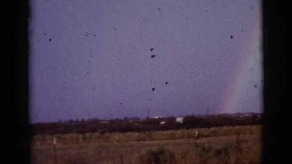 1965 8mm home-movie frame of a rainbow above dry countryside near Tucson, with grasses, scrub, distant poles, and dark frame edges.