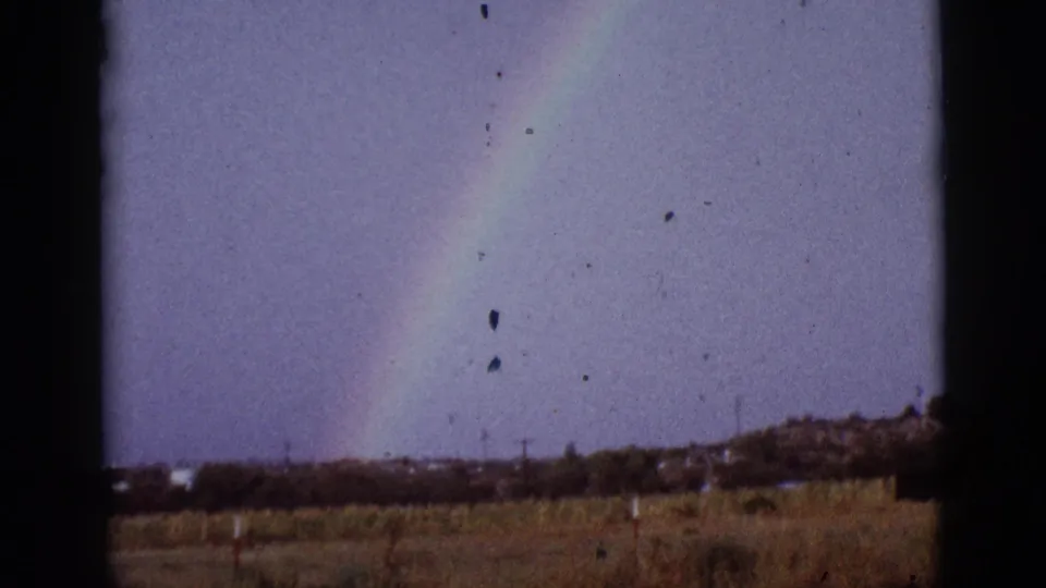A 1965 8mm frame shows a rainbow slanting over dry Tucson-area countryside with low vegetation and visible small-gauge film texture.