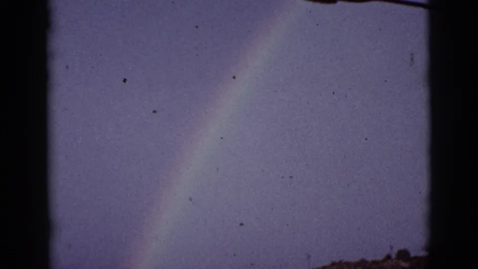 1965 8mm home-movie frame showing a faint rainbow above a rural Arizona field, with distant low land and dark film borders.