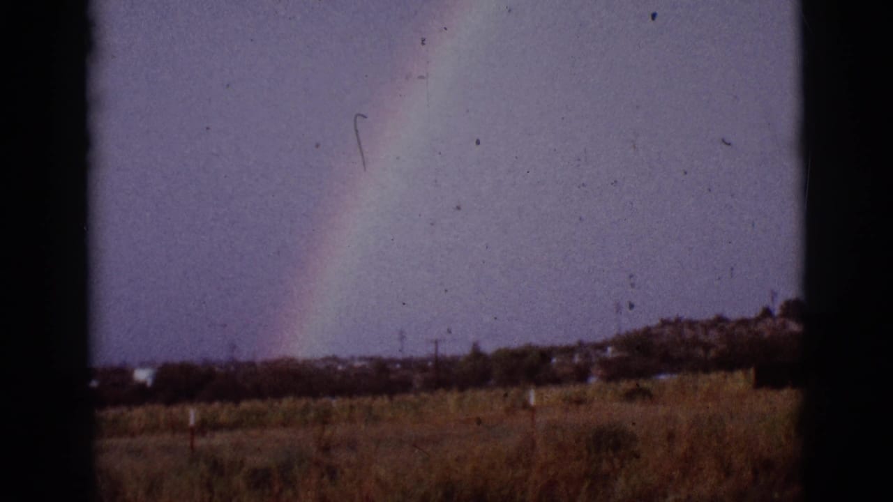 Rainbow over a field with distant trees, structures, and fence posts.