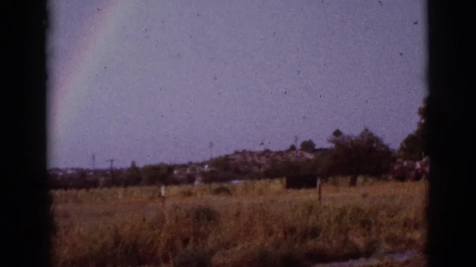 1965 8mm view of a rainbow at the edge of the frame over a grassy rural Tuscon, Arizona landscape with distant trees.