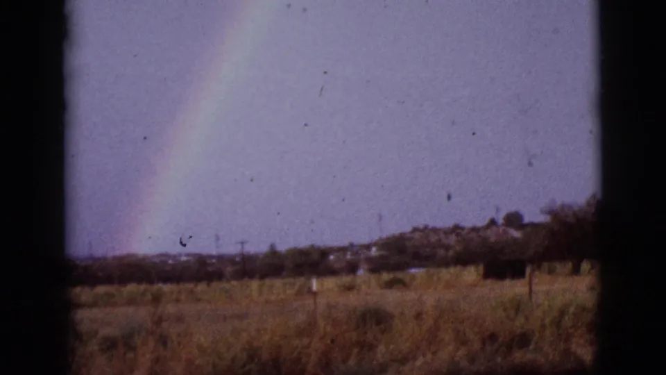 Final 1965 8mm frame showing a faint rainbow over rural Arizona grassland, with trees and low buildings in the distance.