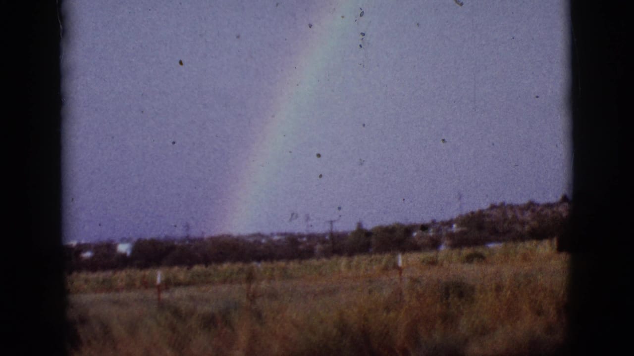 Rainbow rising above a brown field with distant houses and sky.