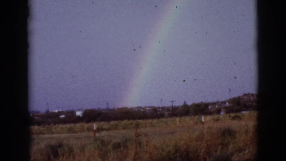 1965 8mm Tucson-area view of a pale rainbow above dry grass and a distant ridge, partly blocked by a dark frame edge.