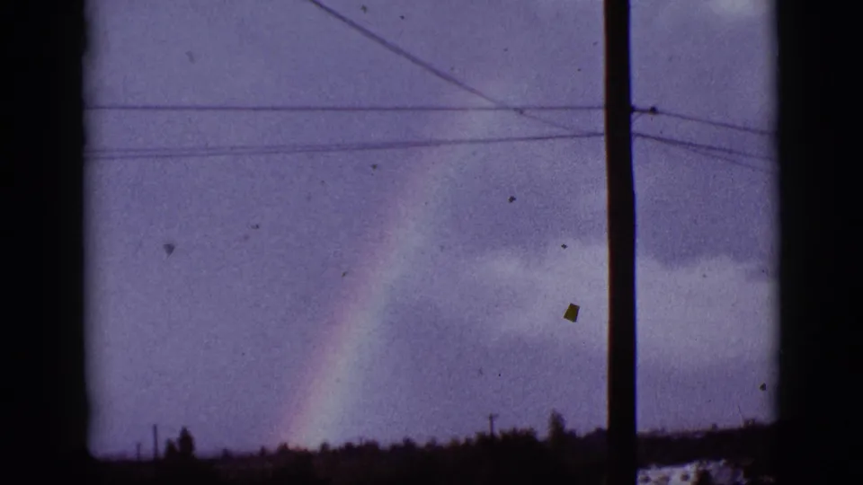 1965 8mm view in Tucson metadata showing a rainbow beyond a window edge, utility pole, power lines, clouds, and low trees.