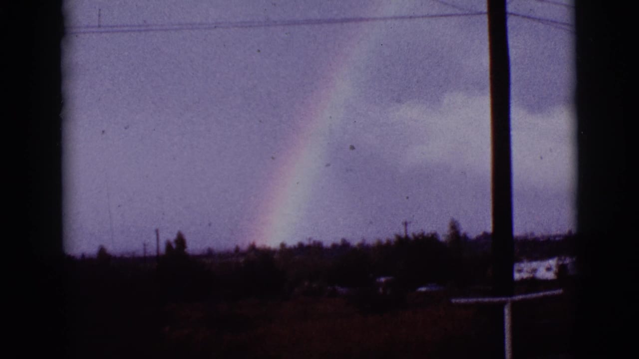 Rainbow segment in cloudy sky with dark vertical framing and distant low structures.