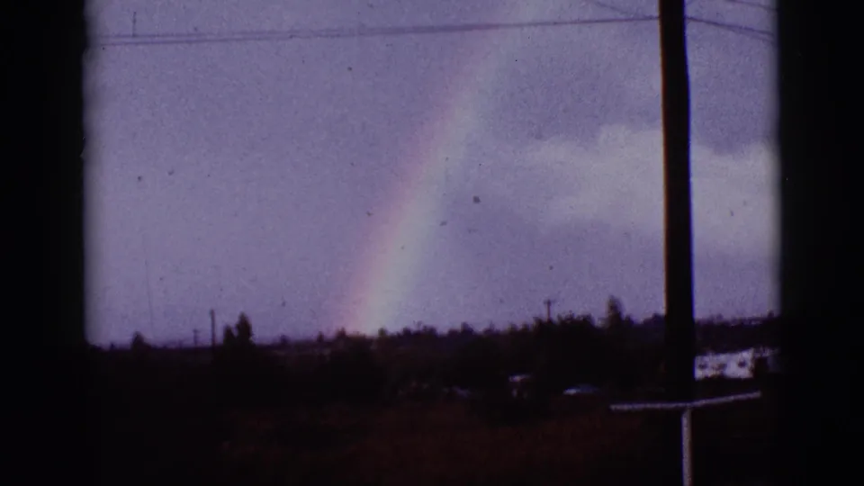 1965 8mm Tucson-area rainbow view framed by dark window edges, with clouds, low vegetation, and small distant structures.