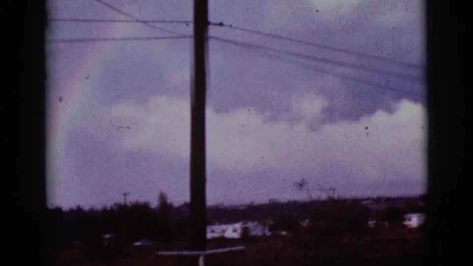 1965 8mm Tucson-area view of a fading rainbow near a utility pole, power lines, clouds, and a low distant horizon.