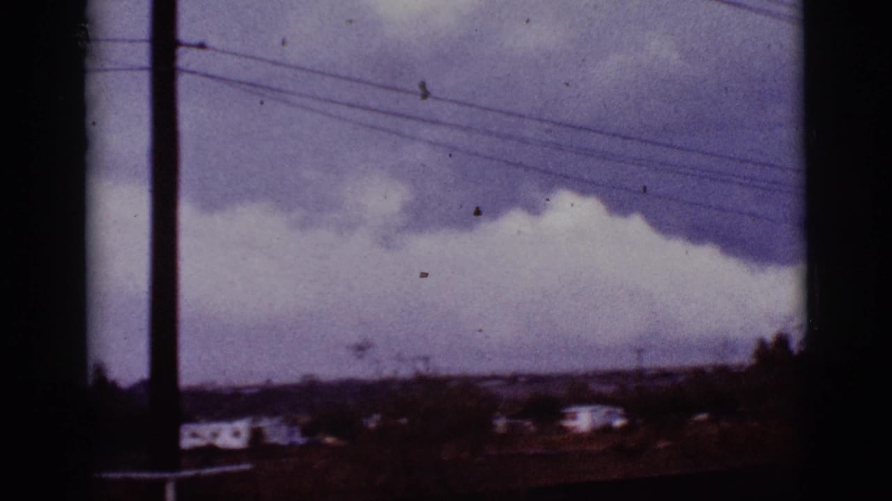 Storm clouds over a low landscape with overhead power lines and distant buildings.