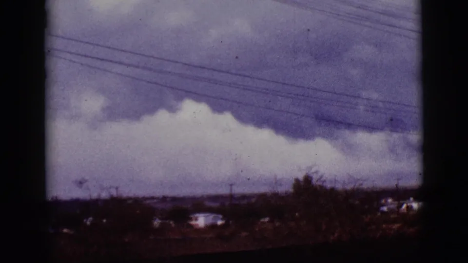 1965 Tucson-area home-movie frame with heavy clouds, power lines, distant homes, and scrub vegetation.