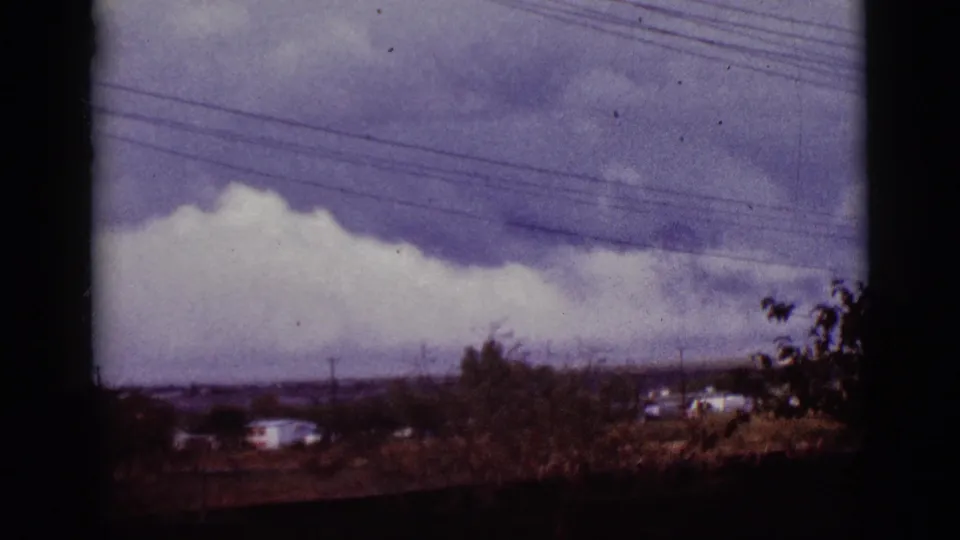 Archival 8mm frame from Tucson, Arizona, showing layered clouds, wires, distant houses, and roadside brush.