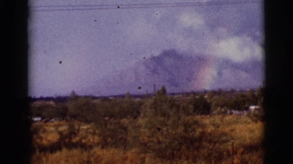 1965 Tucson-area frame with a faint rainbow breaking through clouds above trees, utility poles, and dry terrain.
