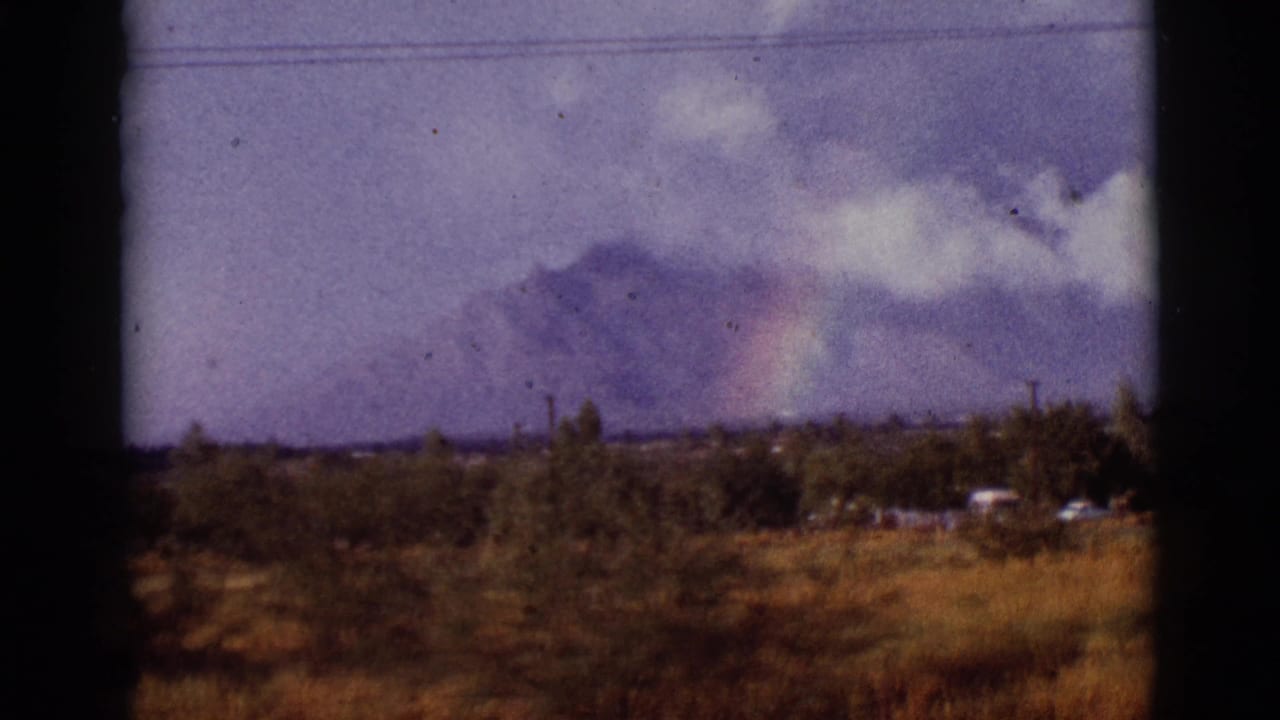 1965 Arizona home-movie frame showing distant mountains, storm clouds, a faint rainbow, dry vegetation, and utility lines near a small settlement.