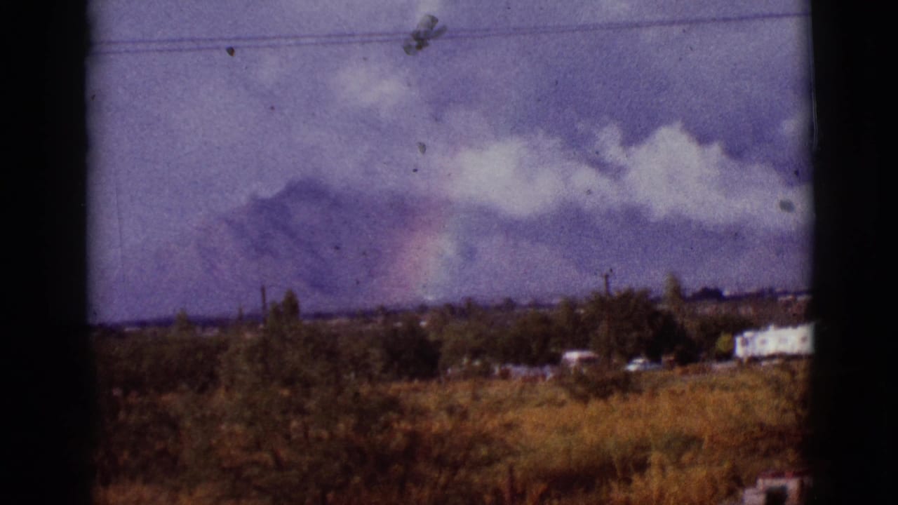 Mountain horizon with clouds, faint rainbow, trees, dry grass, utility lines, and small buildings.