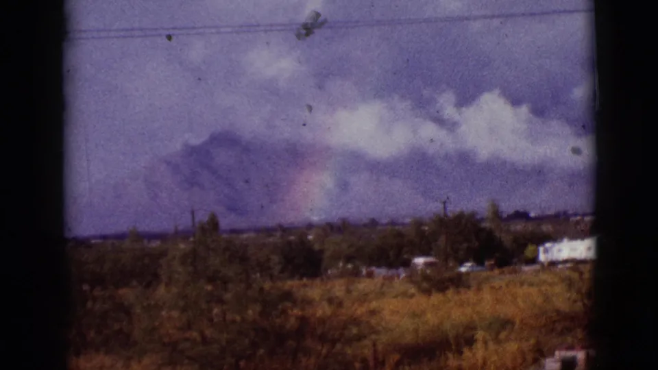 1965 Arizona landscape frame with a faint rainbow against storm clouds over mountains, seen past brush, trees, utility lines, and low buildings.