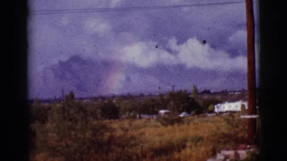 1965 Arizona home-movie frame showing a cloudy mountain backdrop, faint rainbow, dry brush, utility lines, trees, and a dark vertical pole at the right edge.