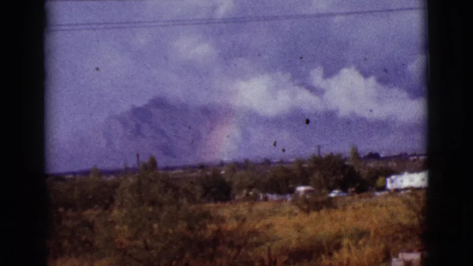 1965 Arizona frame with a faint rainbow and storm clouds over distant mountains, seen across dry grass, trees, utility lines, and small low buildings.