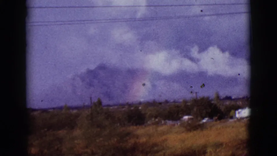 1965 Tucson-area 8mm frame showing a faint rainbow over distant mountains, with dry grass, houses, utility lines, and dark window-like borders.
