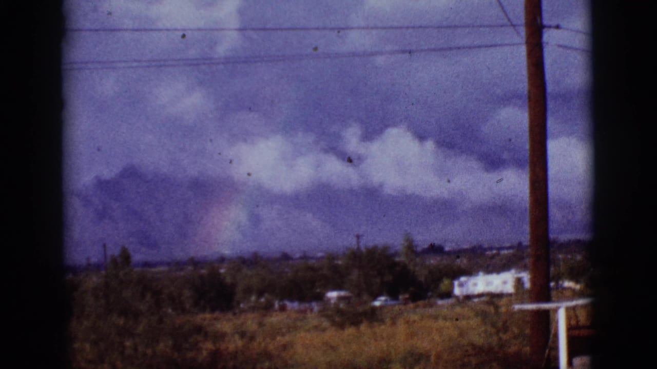 Cloudy mountain horizon with a faint rainbow, small buildings, utility pole, and dark vertical framing.