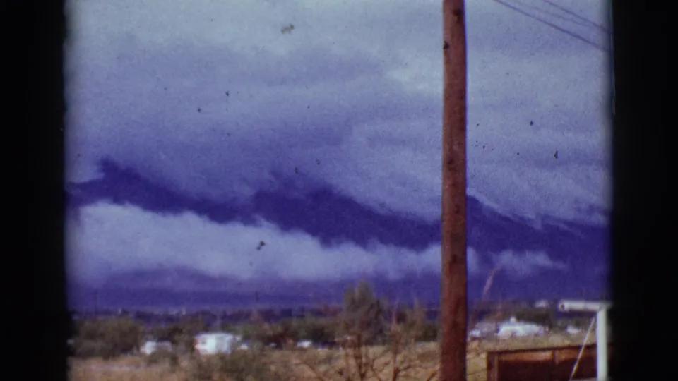 1965 Tucson-area 8mm frame with heavy purple storm clouds over mountains, foreground houses and dry grass, plus a utility pole near the center.