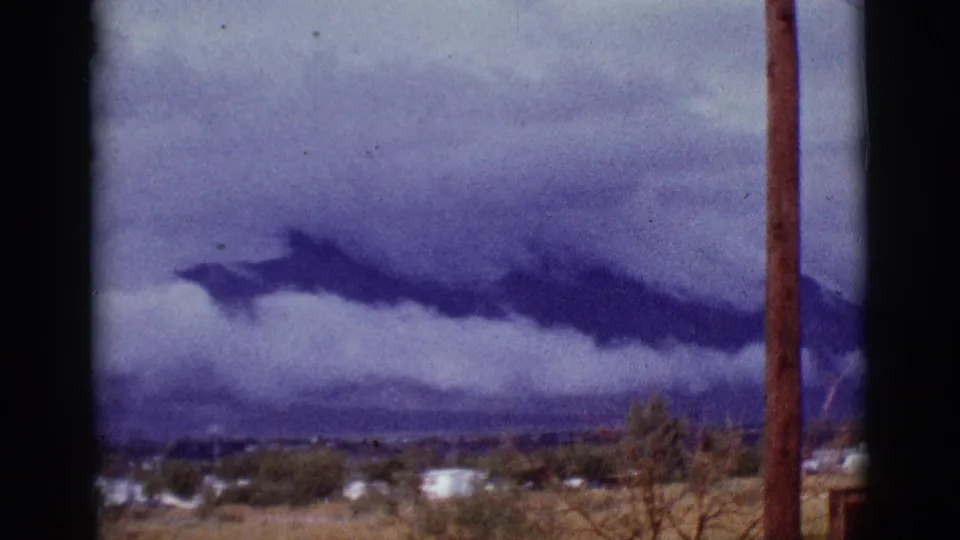 1965 Tucson-area home-movie frame showing a dark cloud mass over distant mountains, with scattered houses, trees, dry grass, and a pole at right.