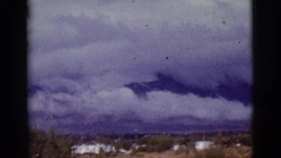 1965 Tucson-area home-movie frame showing a dark cloud band above distant mountains, low trailers and desert vegetation.