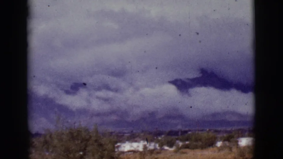 Soft 8mm frame from 1965 showing storm clouds over a Tucson-area desert scene with trailers and scrub in the foreground.