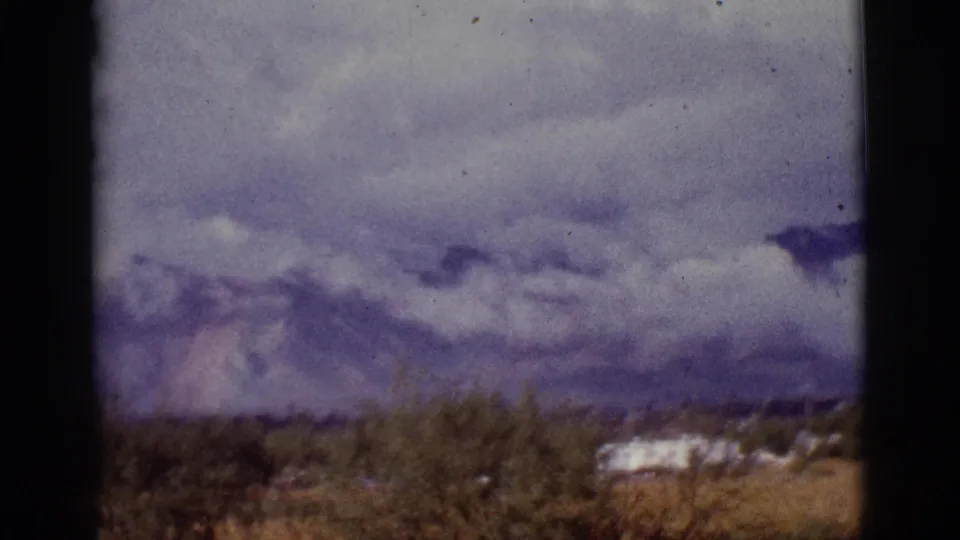 1965 8mm Tucson-area frame with desert scrub, white trailers, distant mountains and heavy clouds under visible film borders.