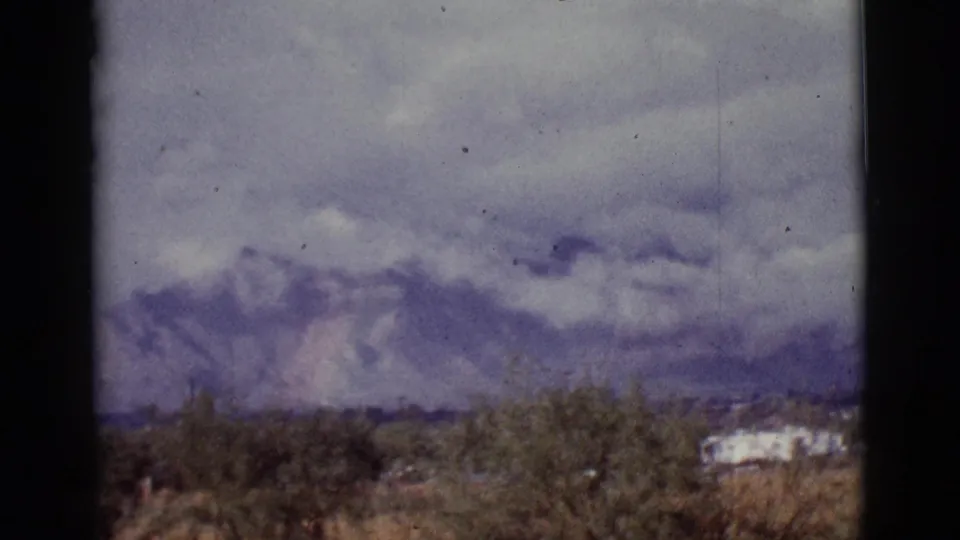 1965 Tucson-area 8mm frame showing desert shrubs, low buildings, and purple-gray mountains beneath heavy clouds.