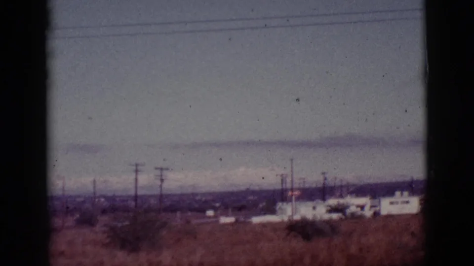 1965 8mm view of Tucson-area scrubland with utility poles, white roadside buildings, and muted mountains under a gray sky.