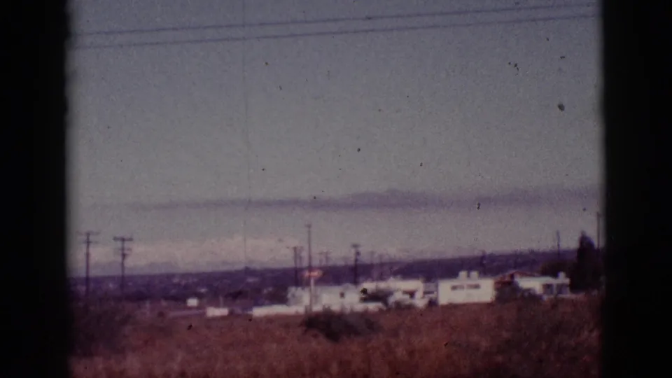 Tucson-area home-movie frame showing low buildings, utility poles, dry brush, and distant mountains under a subdued sky.