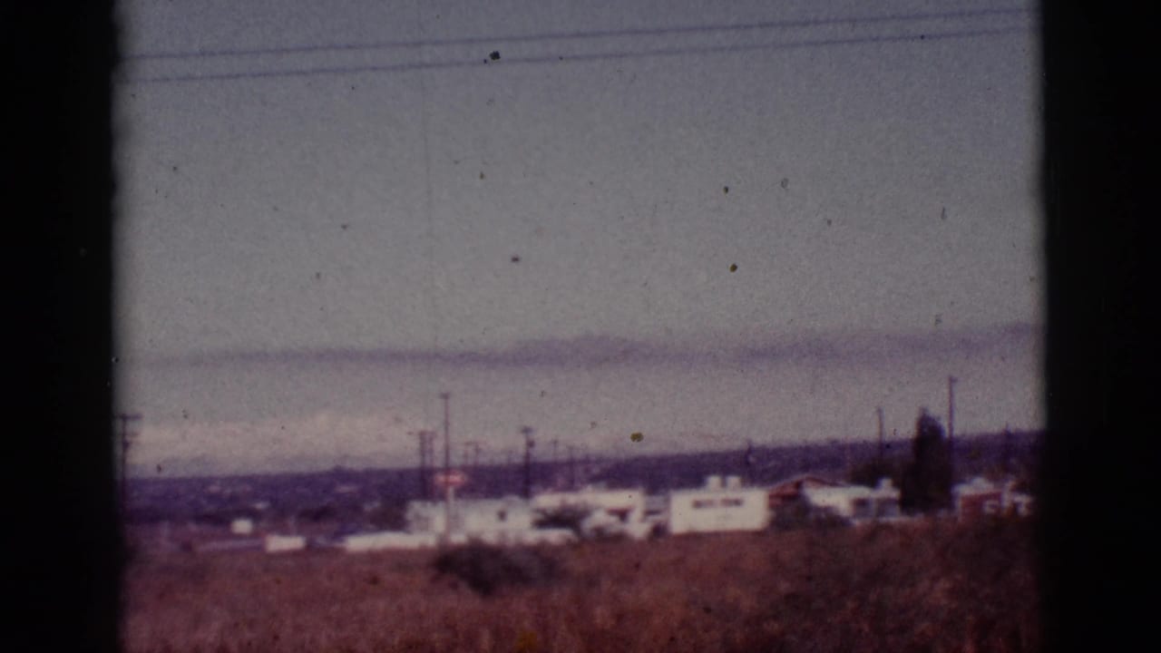 Distant white mobile homes beyond dry desert brush with utility poles and hazy hills.