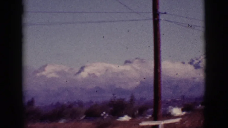 A 1965 Tucson-area view shifts toward distant snow-capped mountains beyond roadside brush, wires, and a tall utility pole.