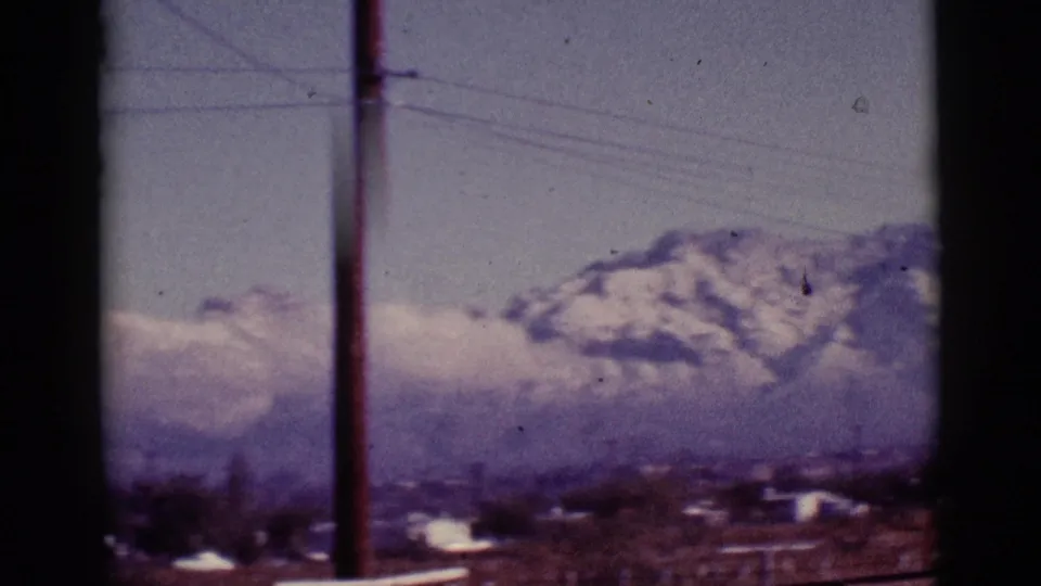 1965 8mm Tucson-area view of distant snow-capped mountains with utility lines, low buildings, haze, and dark framing edges.