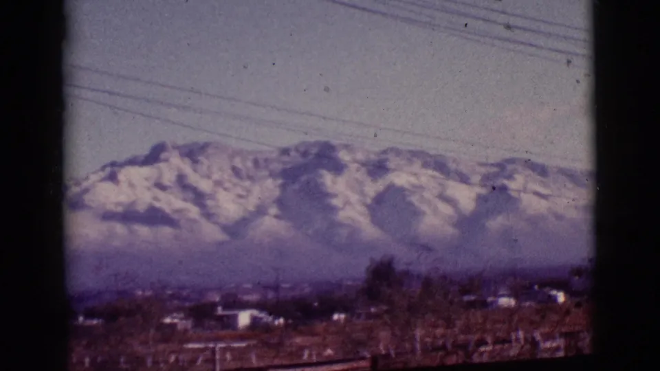 1965 8mm view toward Tucson-area snow-capped mountains, with overhead wires, low structures, haze, and dark film or window borders.