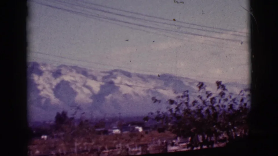 1965 Tucson-area mountain view with snowy ridges, overhead power lines, sparse trees, low buildings, and soft 8mm color shift.