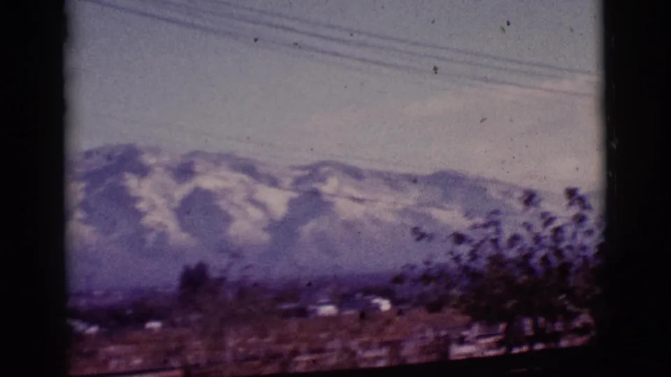 1965 Tucson-area 8mm frame showing distant snow-capped mountains, sparse vegetation, low development, overhead wires, and haze.