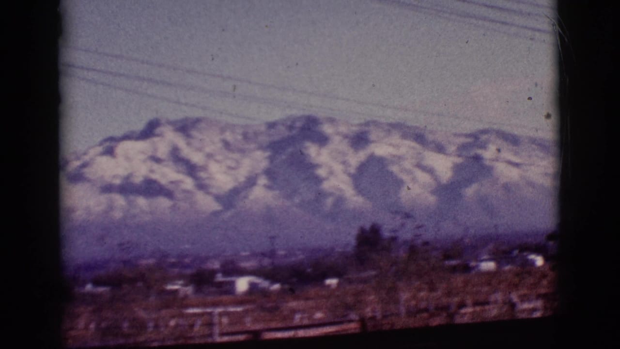 1965 8mm view near Tucson showing snow-capped mountains behind a brown field, distant structures, trees and overhead power lines.