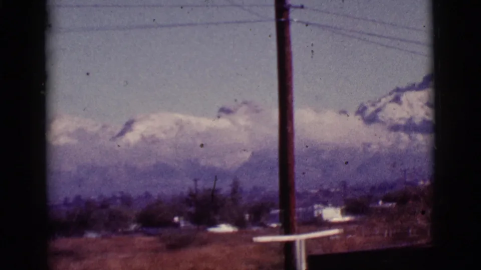 1965 8mm frame from Tucson showing a roadside utility pole against snowy mountain peaks, clouds and a dry field.