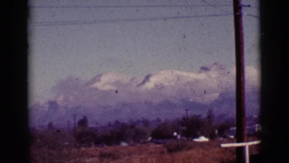 1965 Tucson-area landscape with snow-covered mountains partly veiled by clouds beyond a dry field and scattered trees.
