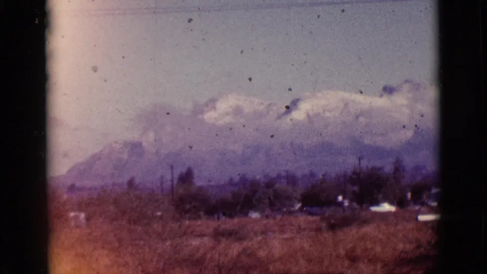 Vintage 8mm frame from 1965 near Tuscon, Arizona, with snow-capped mountains seen past dry grass, brush, and faint roadside structures.