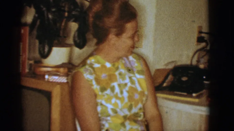 1966 Clarkdale home-movie frame showing a woman in a yellow-green floral dress near a TV console and telephone.
