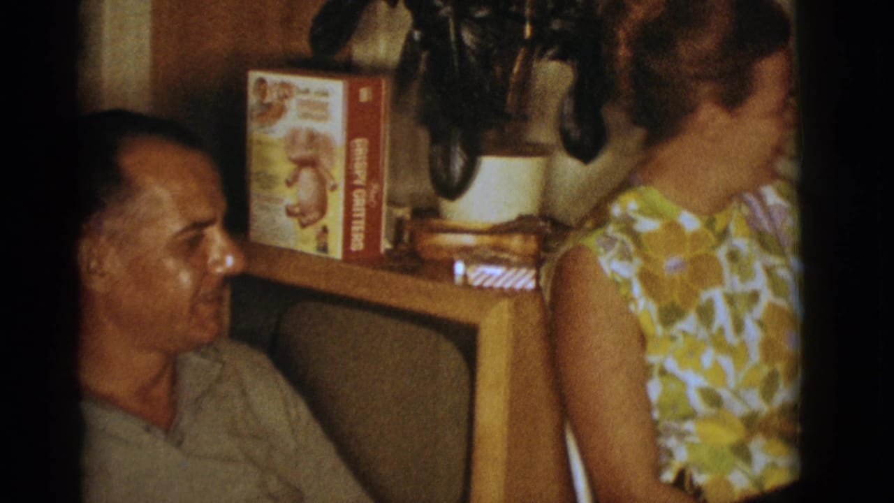 Seated man in a light polo shirt looks toward a woman in a living room.