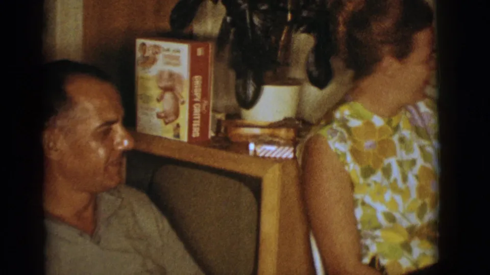 1966 Clarkdale home-movie frame with a seated man looking toward a woman beside a console with a cereal box and plant.