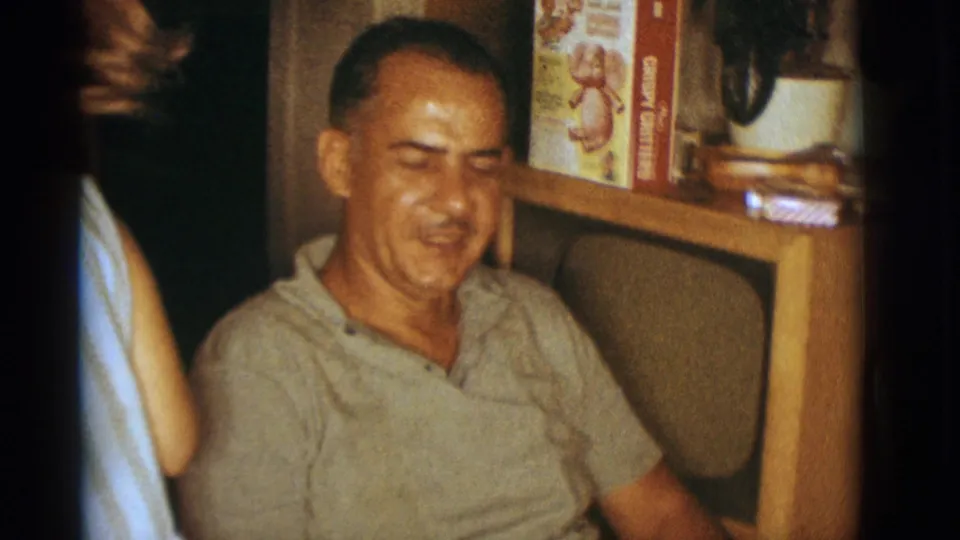1966 Clarkdale home-movie frame of a smiling seated man in a private living room with a console, plant, and cereal box behind him.