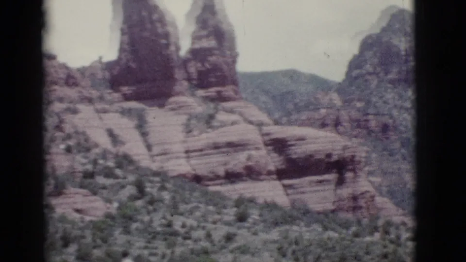1966 home-movie frame near Clarkdale showing vertical red rock spires, banded sandstone ledges, canyon slopes, and desert plants.