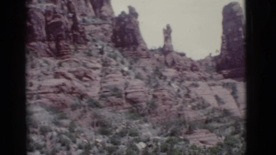 Late 1966 8mm frame near Clarkdale with rugged red rock slope, upright formations, layered sandstone, scrub vegetation, and gate-edge shadow.